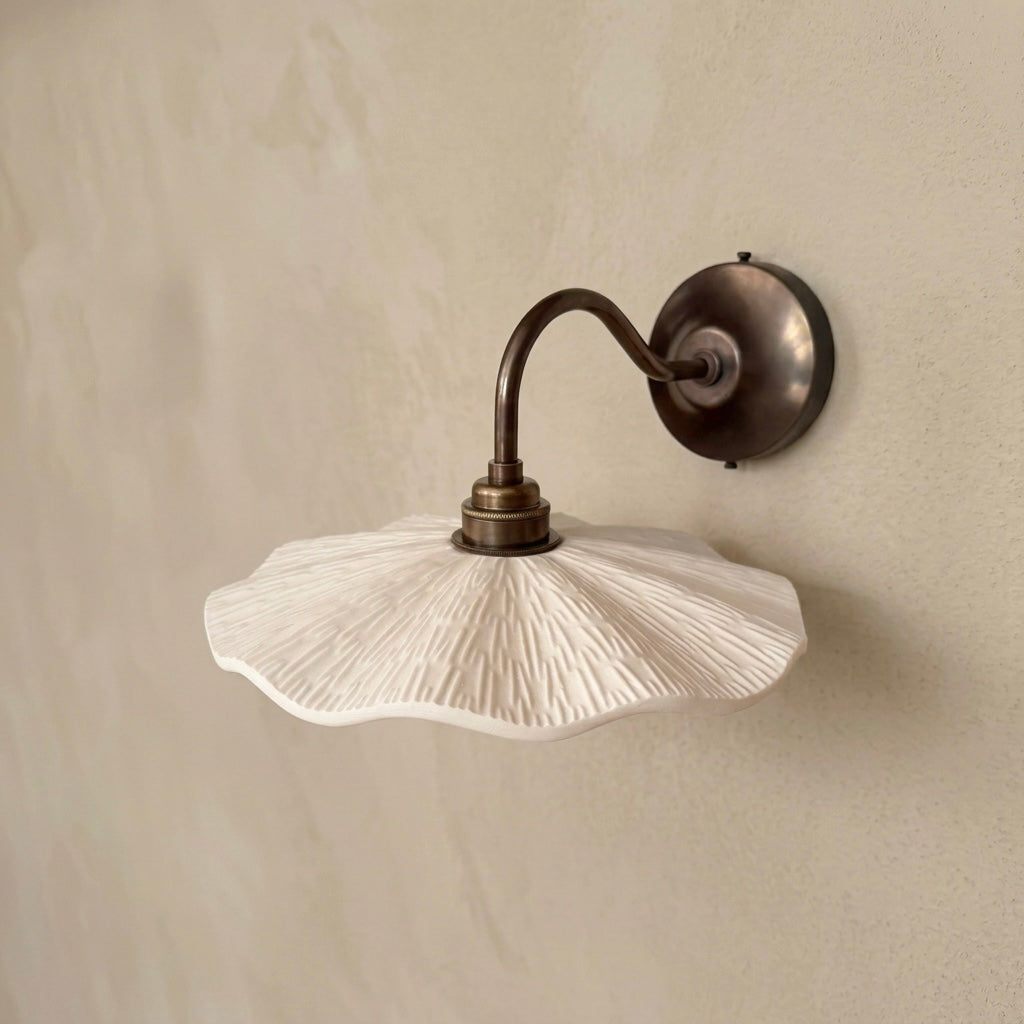 A wall-mounted sconce with a textured white scalloped shade and an antique bronze curved arm and circular wall mount, shown against a warm beige plaster wall.