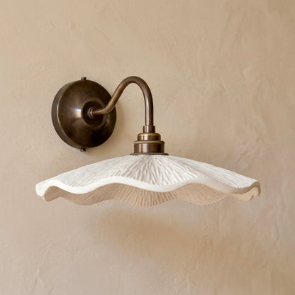 A wall-mounted sconce with a textured white wavy shade and an antique bronze curved arm and wall plate, shown against a warm beige plaster wall.