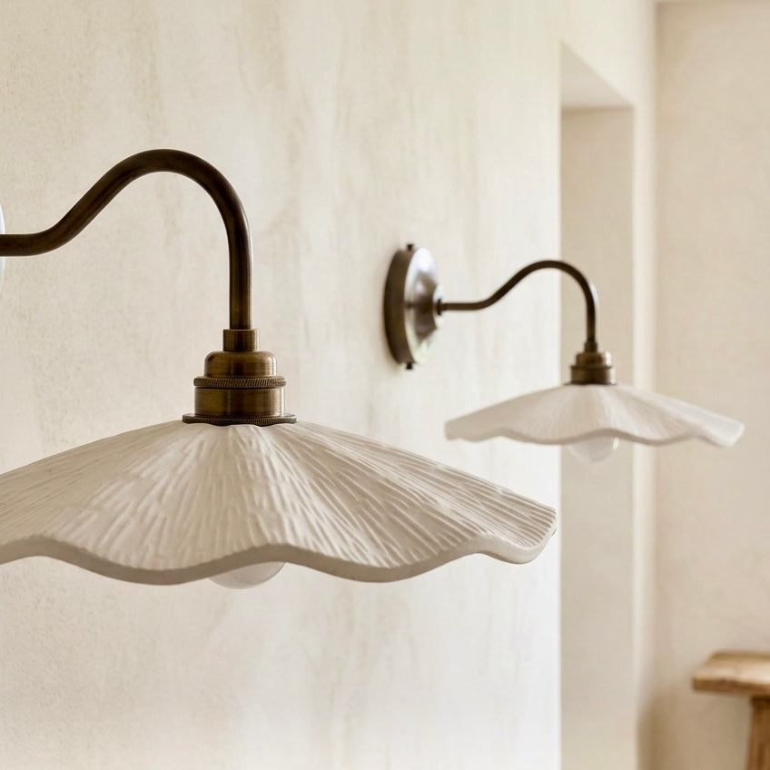 A close-up of a wall-mounted sconce with a textured white fluted shade and an antique brass curved arm, shown against a warm beige plaster wall. A second matching sconce is visible in the background.