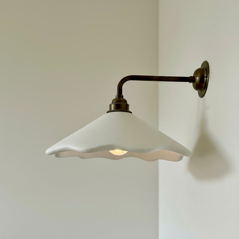 Wall-mounted light fixture with white shade and brass arm on a plain wall.
