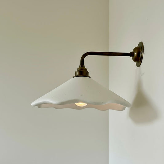 Wall-mounted light fixture with white shade and brass arm on a plain wall.