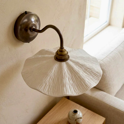A wall-mounted sconce with a large textured white pleated shade and an antique brass curved arm and wall plate, shown installed on a beige wall beside a sofa and window.