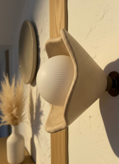 Close-up of a sculptural beige wall light bulb on a brass fixture against a neutral wall.