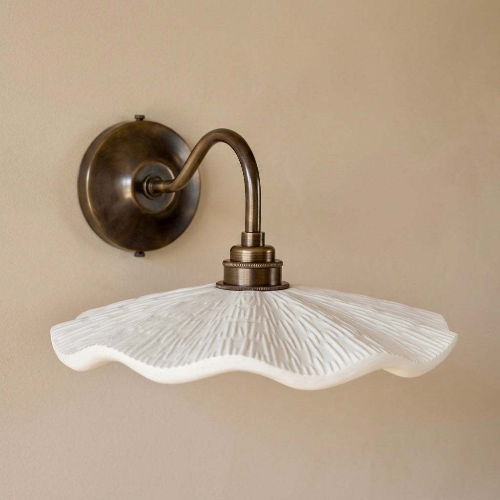 A wall-mounted light fixture with a textured white scalloped glass shade and an antique bronze metal wall plate and curved arm, shown against a beige wall.