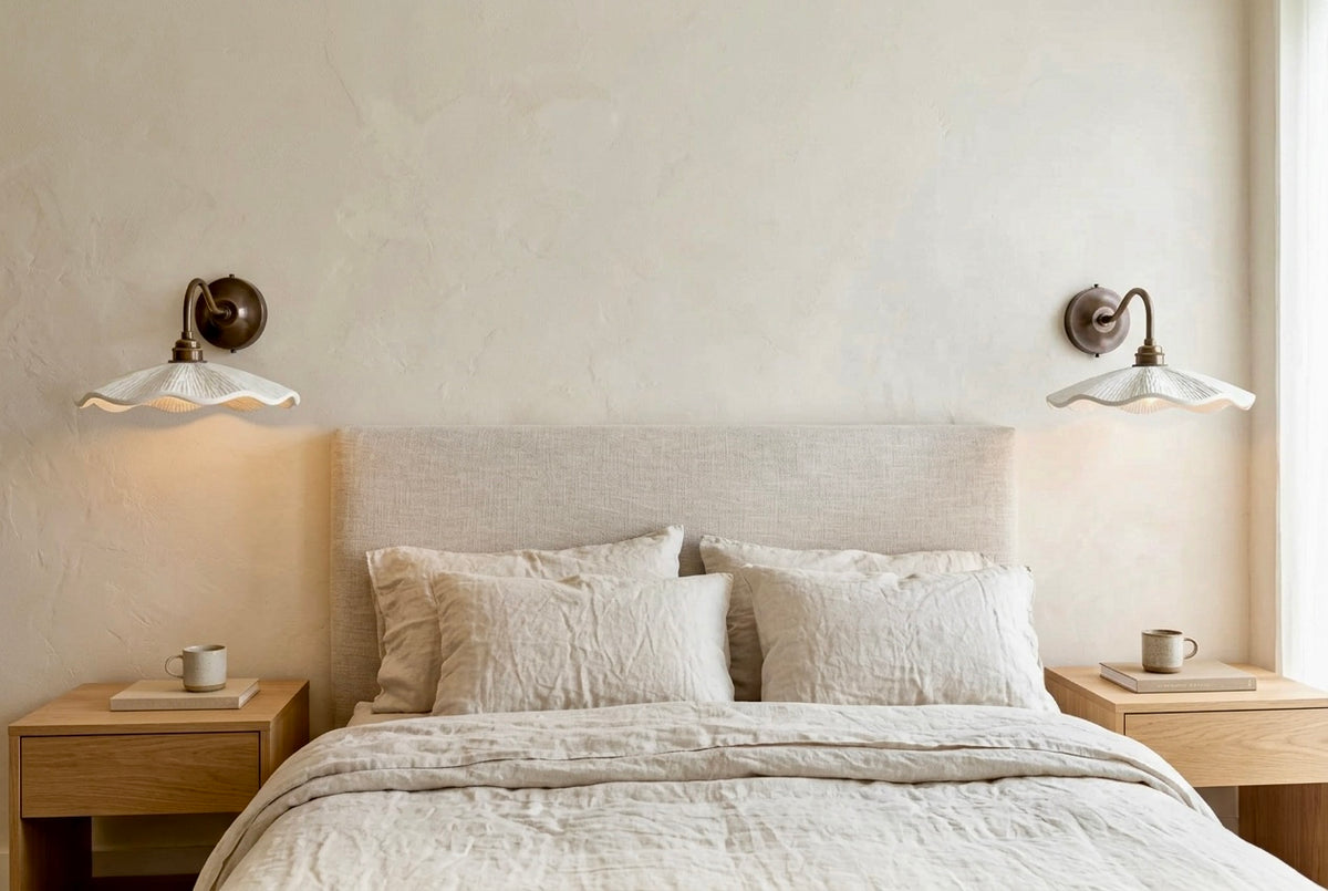 A minimalist bedroom scene featuring a neatly made bed with textured off-white bedding and pillows, a light beige upholstered headboard, two matching wooden nightstands with mugs, and two wall-mounted sconces on a textured cream wall.