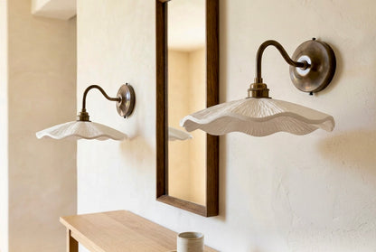 A pair of wall-mounted sconces with wavy white glass shades and dark metal arms, shown installed on a textured cream wall beside a wooden-framed mirror and a light wood console table.