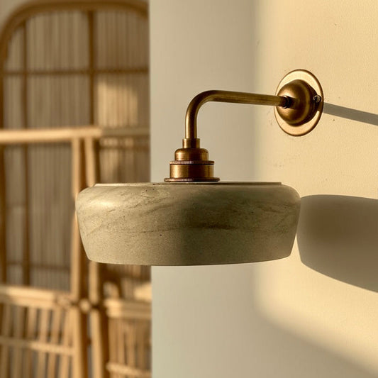 Wall-mounted light fixture with concrete base and brass arm in a softly lit room.