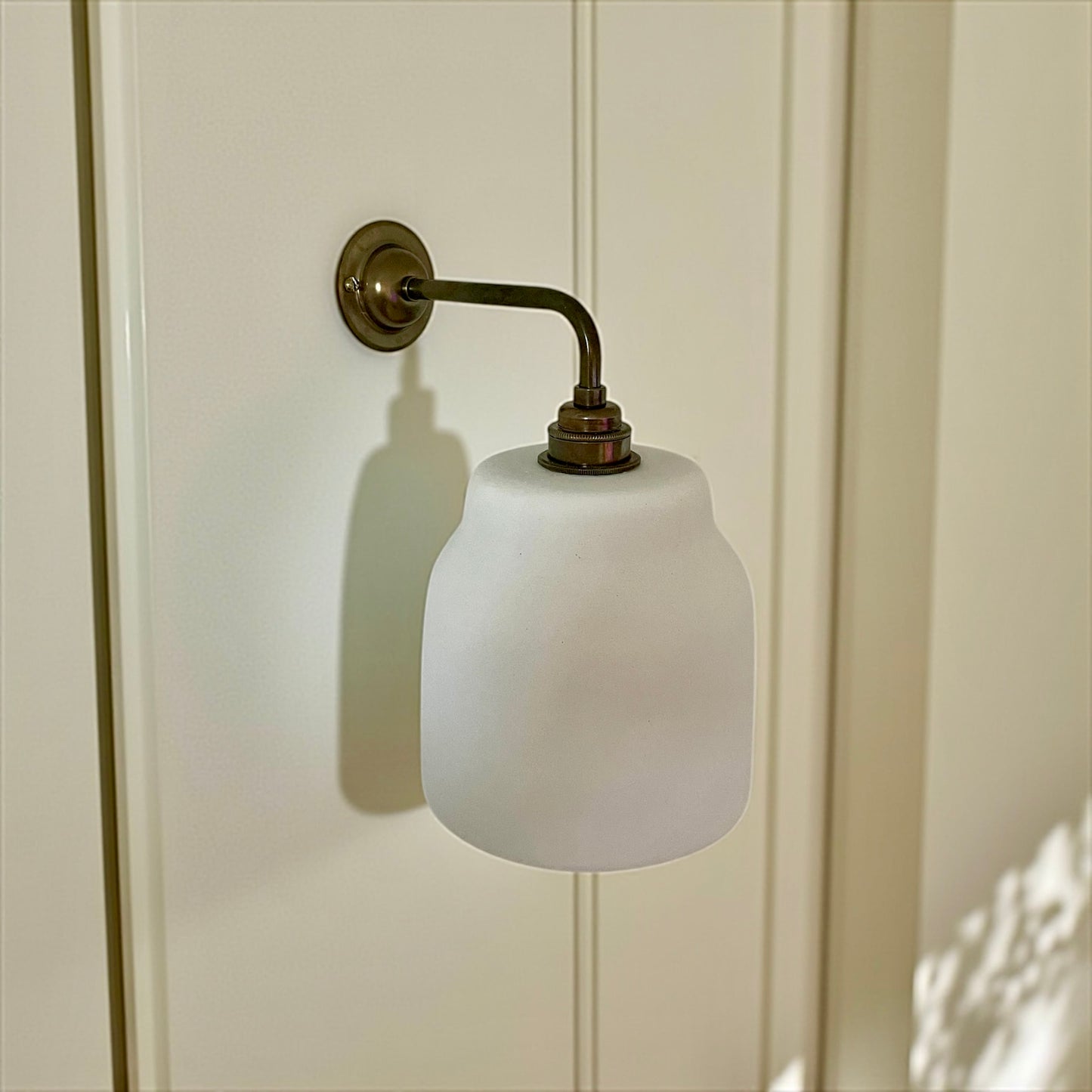 Wall-mounted light fixture with white glass shade and brass fixture on a white paneled wall.