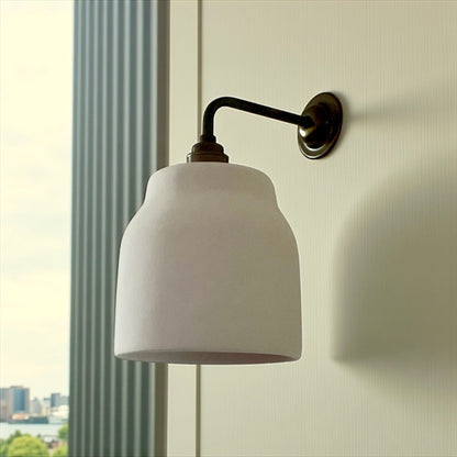 Wall-mounted lamp with white shade and brass arm against a light wall.