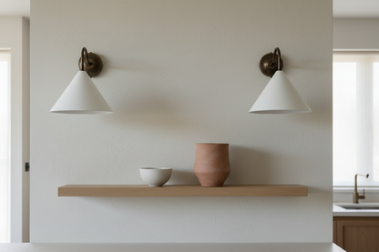 Two wall-mounted lamps with white shades on a plain wall above a wooden shelf with ceramic items.